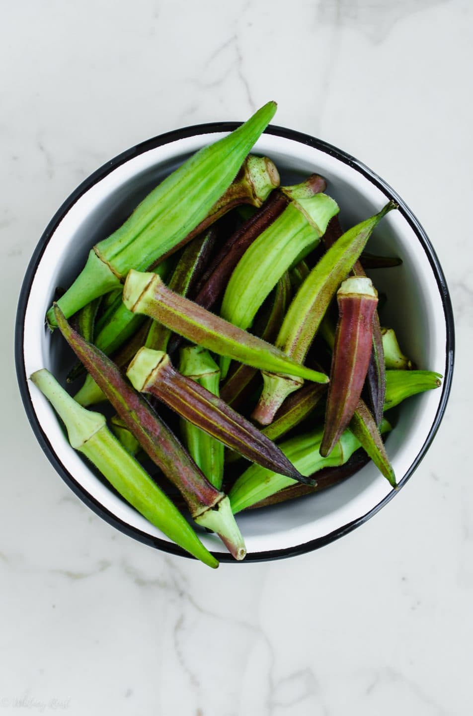 A white bowl filled with fresh okra pods on a white marble surface.