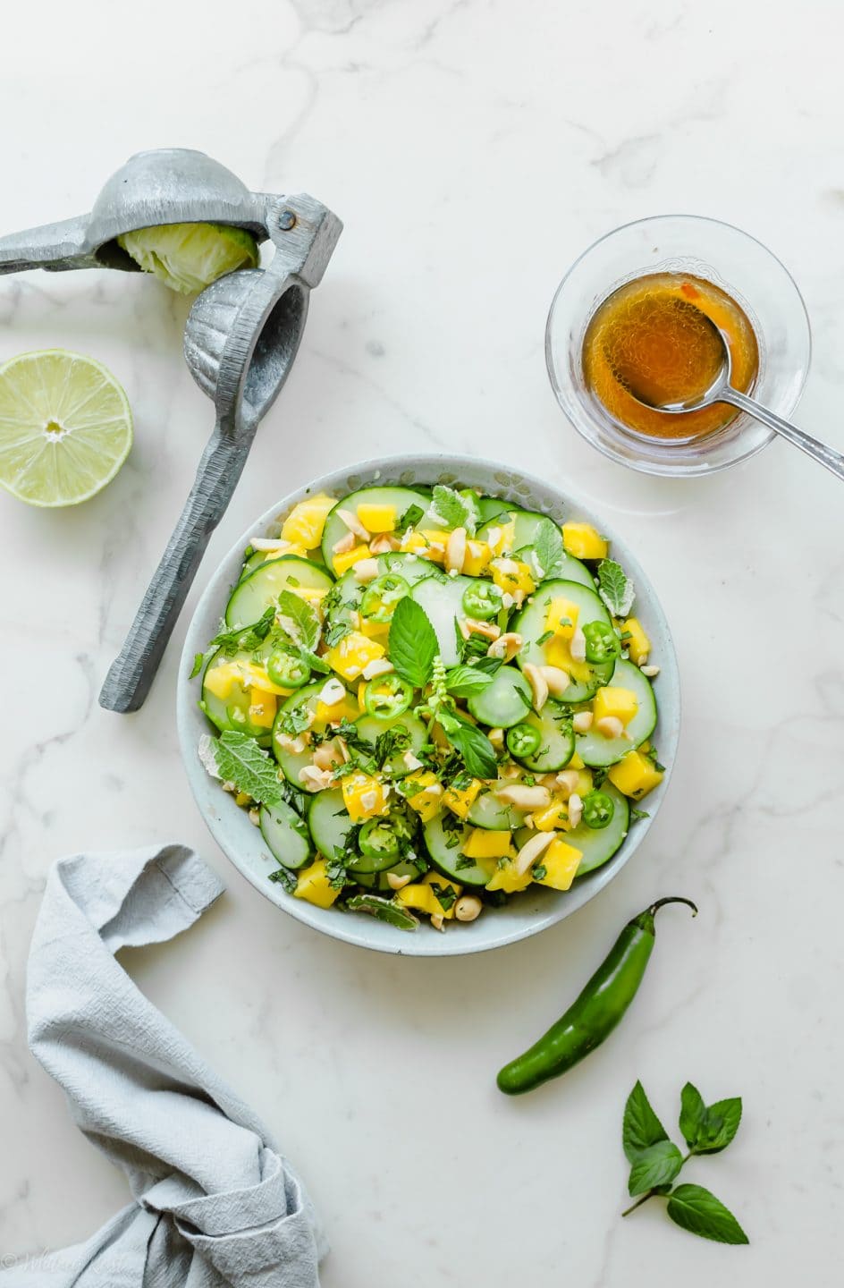 A grey bowl with Thai cucumber mango salad with a small dish of vinaigrette and fresh limes on the side.