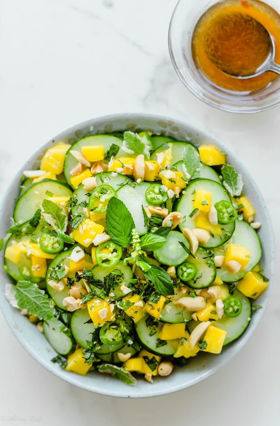 A close up shot of a bowl of Thai cucumber mango salad with a bowl of vinaigrette on the side.