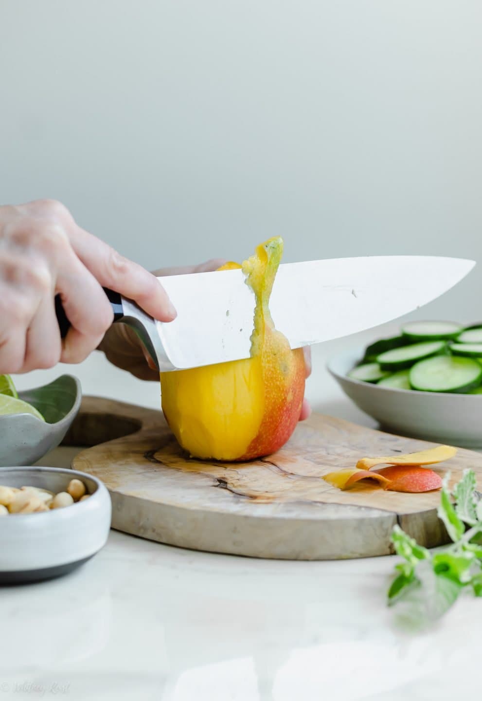 A hand and knife peeling the skin off of a mango on a cutting board. 
