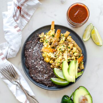 A grey plate with migas and refired black beans with a fork and jar of taco sauce on the side.