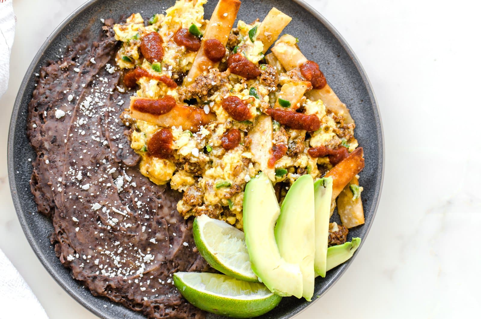 A grey plate with migas and refired black beans with a fork and jar of taco sauce on the side. 