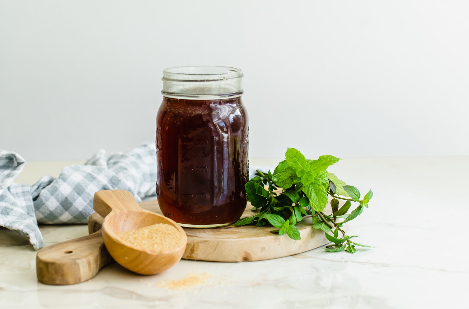A jar of fresh mint simple syrup sitting on a wooden cheese board with a scoop of raw sugar and a bunch of fresh mint on the sides.