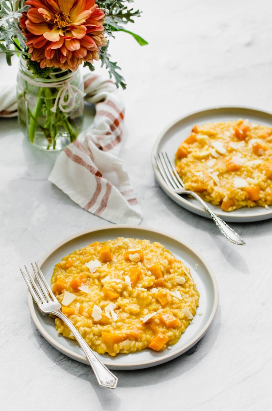Two plates with butternut squash risotto on a white background with a vase of flowers on the side. 