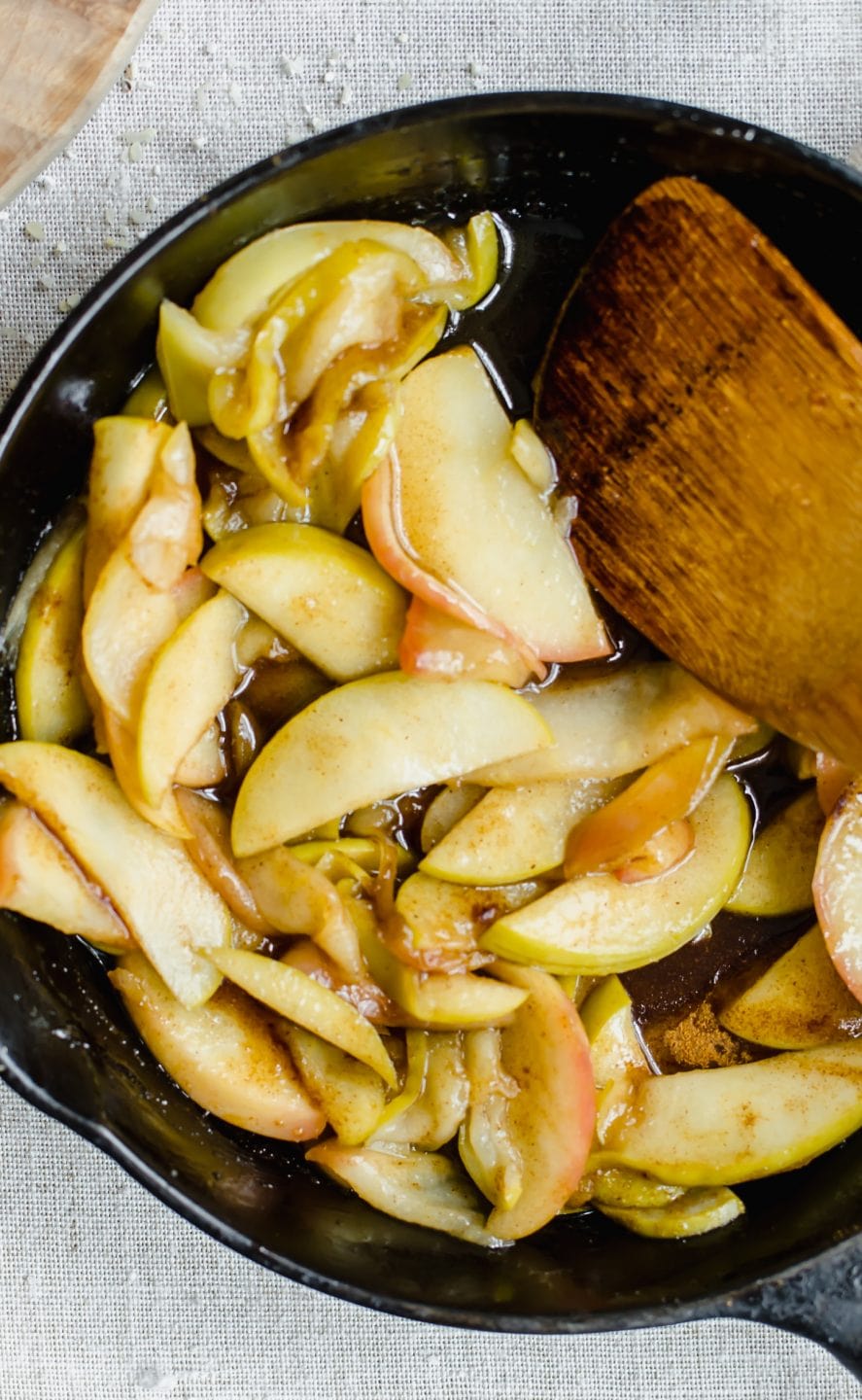A cast iron skillet with sauteed cinnamon apples and a wooden spoon.