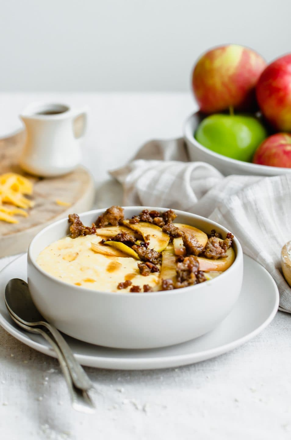 A grey bowl filled with cheddar grits, cinnamon apples, and sausage crumbles with a plate of apples in the back.