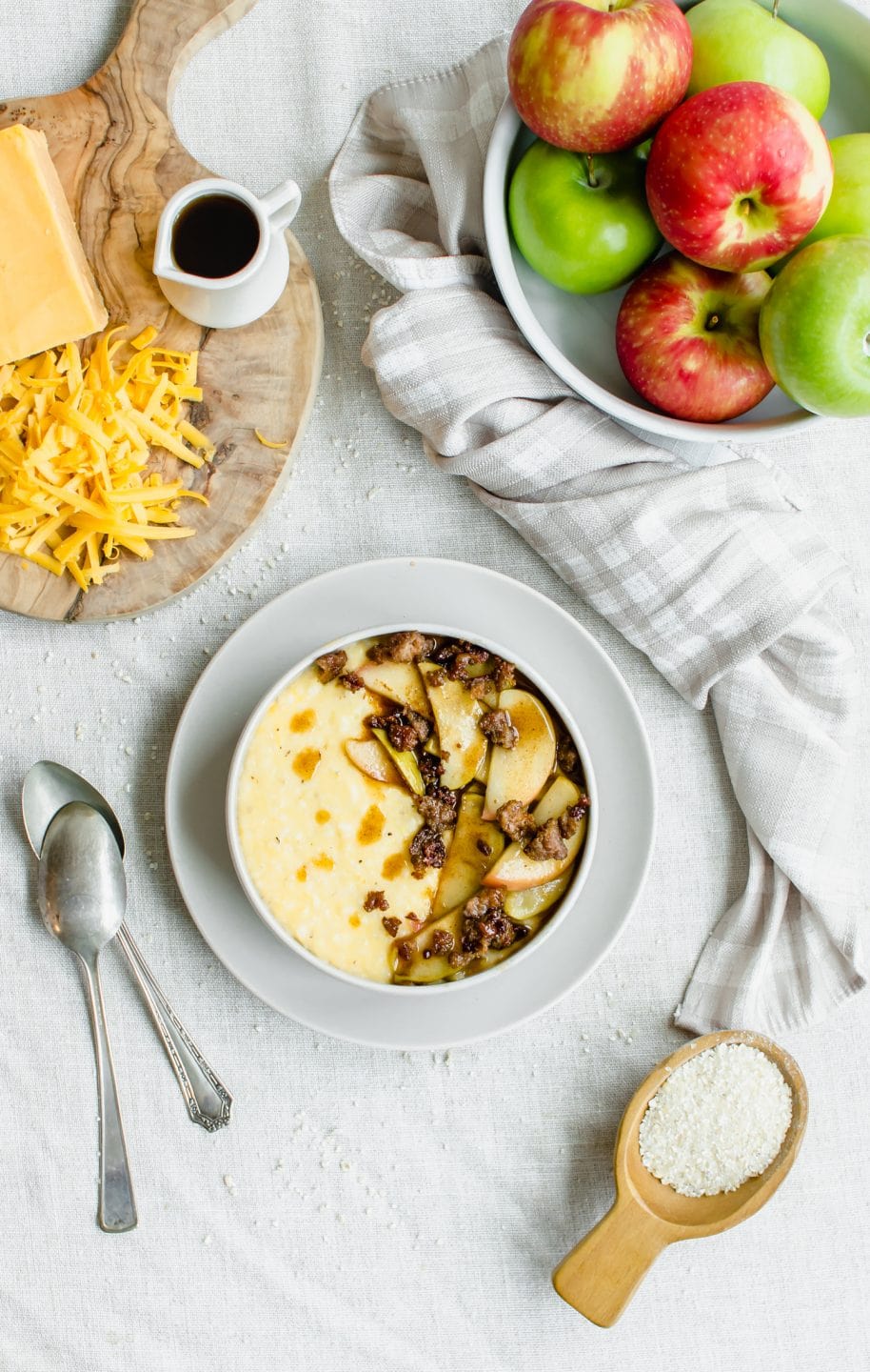 An overhead shot of a bowl of grits topped with cinnamon apples and turkey sausage with ingredient prep bowls on the side.