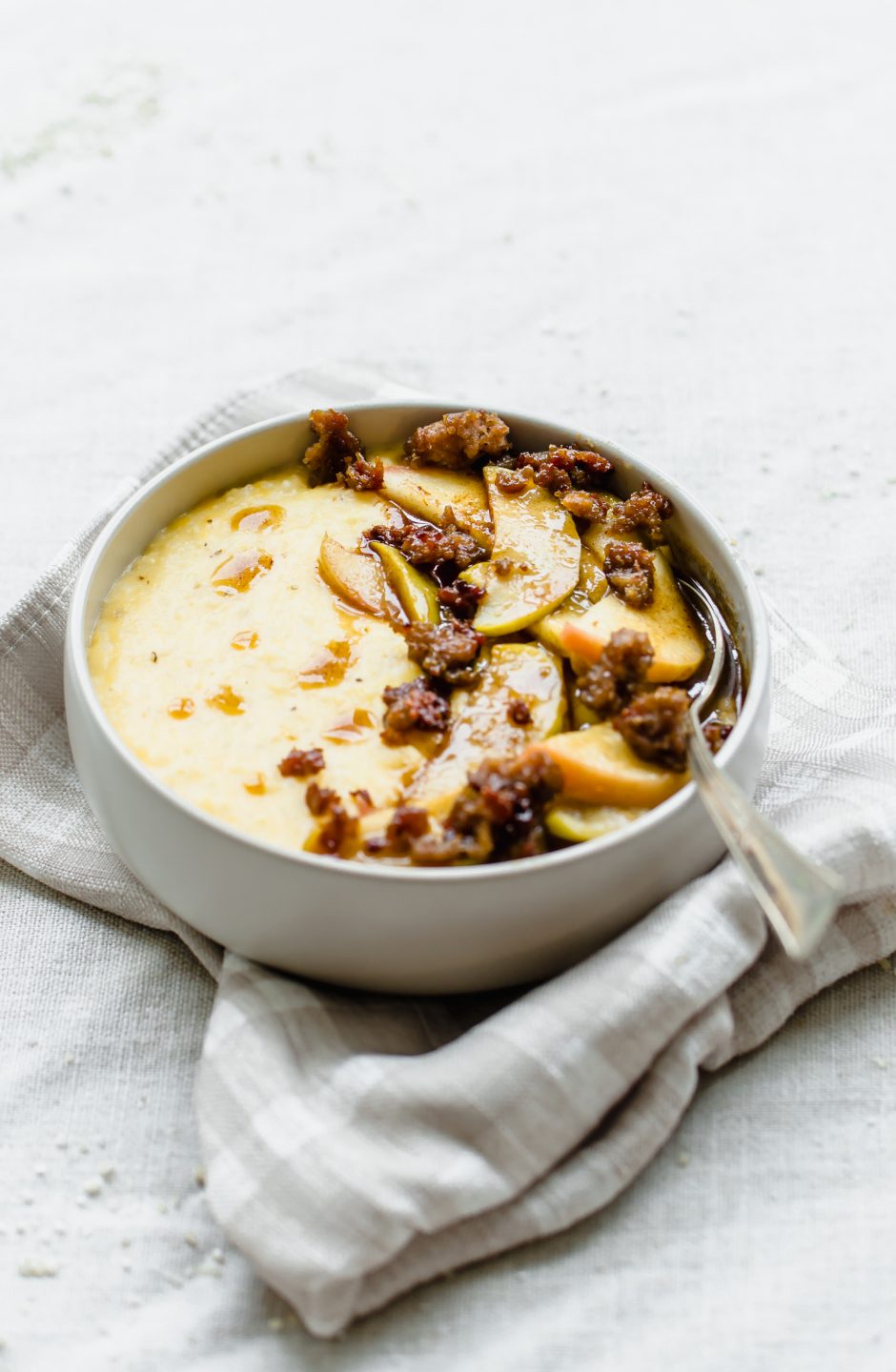 A white bowl filled with grits and cinnamon apples sitting on a taupe and cream gingham napkin.