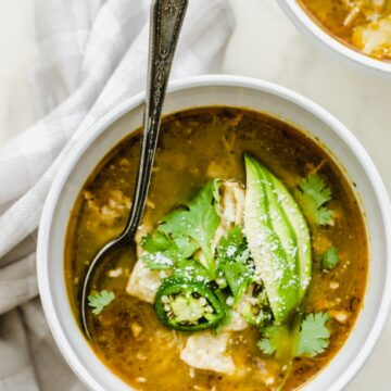 A white bowl of soup filled with posole verde and garnished with avocado and cilantro.