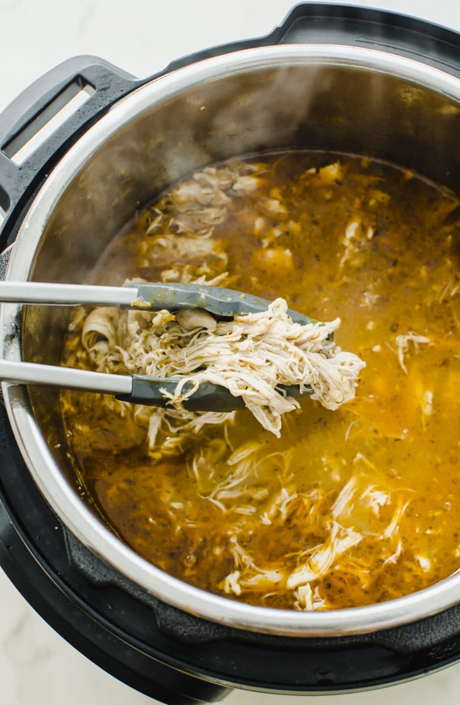 Tongs holding shredded chicken coming up from a soup inside an Instant Pot.