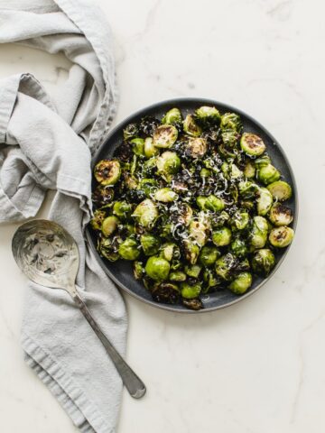 An overhead shot of a plate of roasted Brussels sprouts with a grey linen towel and serving spoon on the side.