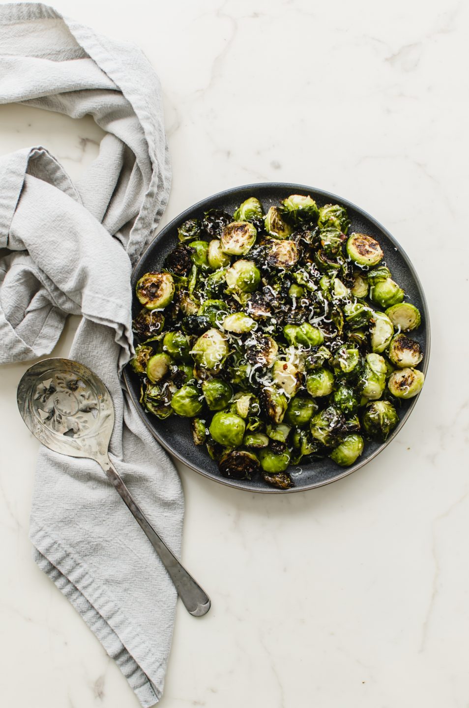 An overhead shot of a plate of crispy roasted Brussels sprouts with a grey linen towel and serving spoon on the side.