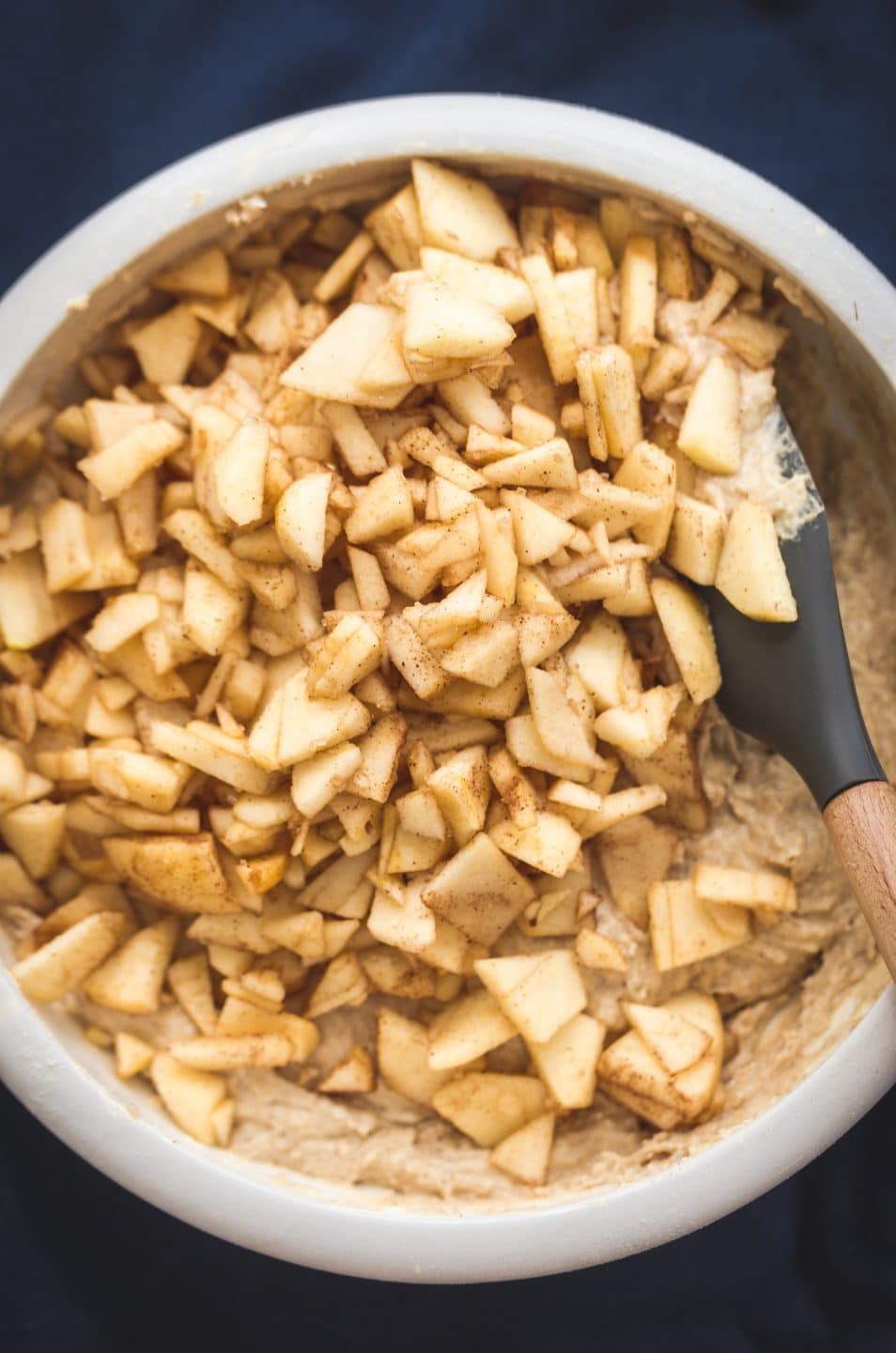 A white mixing bowl filled with cake batter and cinnamon apples being folded in.