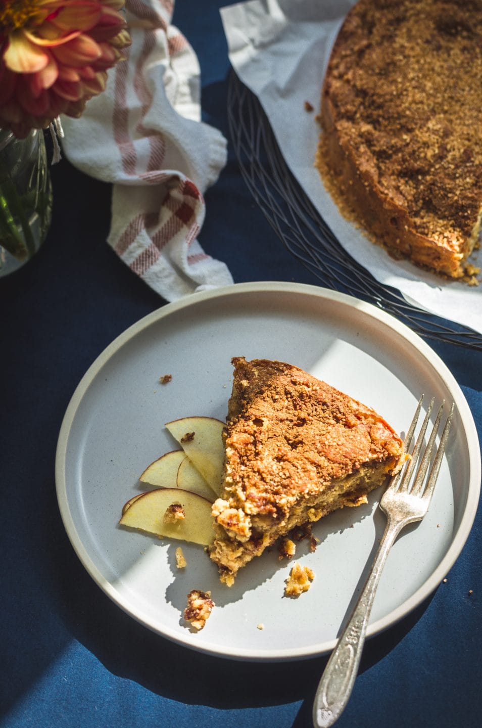 A slice of apple cake on a blue plate with thinly sliced apple and a fork on the side.