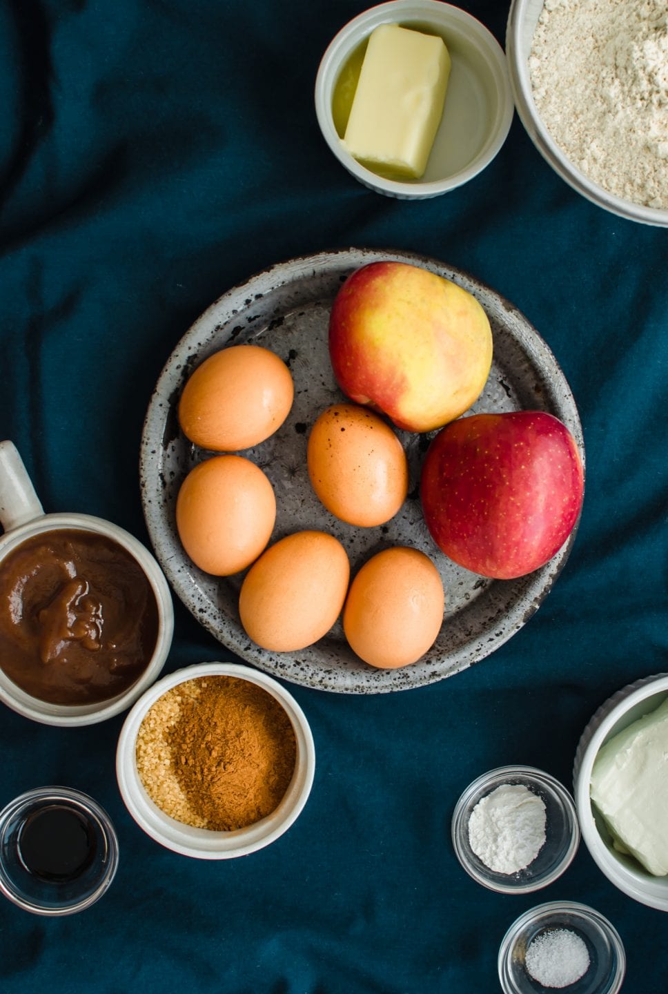 A blue plate with apples and eggs surrounded with small prep bowls with apple cake ingredients.