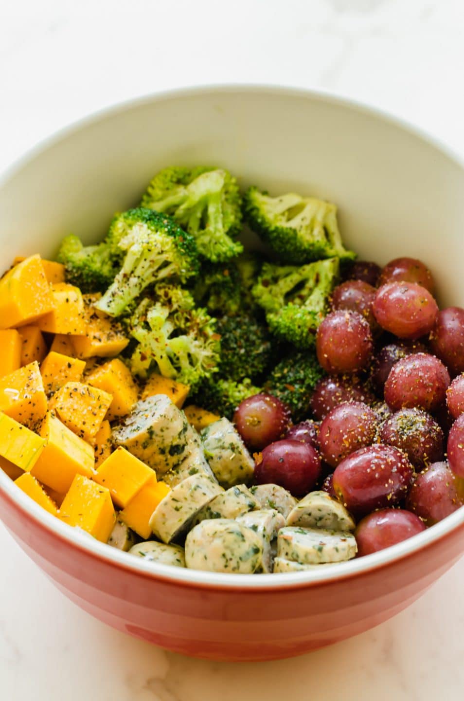 A large bowl filled with cut broccoli florets, cubed butternut squash, red grapes, and chicken sausage.