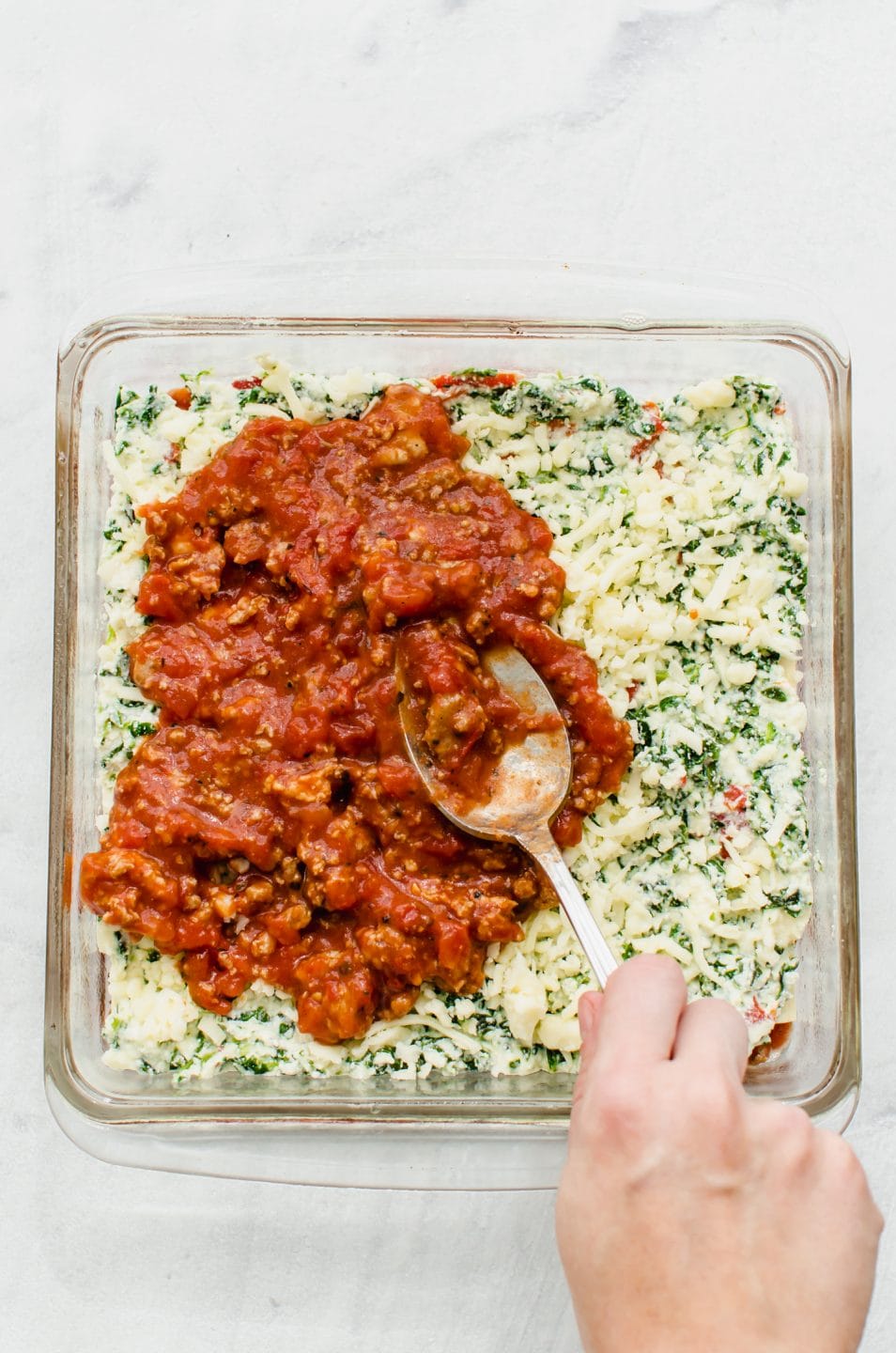 A spoon spreading meat sauce on top of the cheese layer of a lasagna.