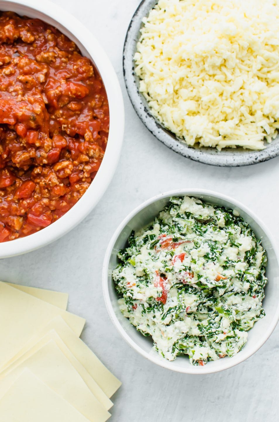 Prep bowls with ingredients for making Turkey Florentine Lasagna on a white stone background.