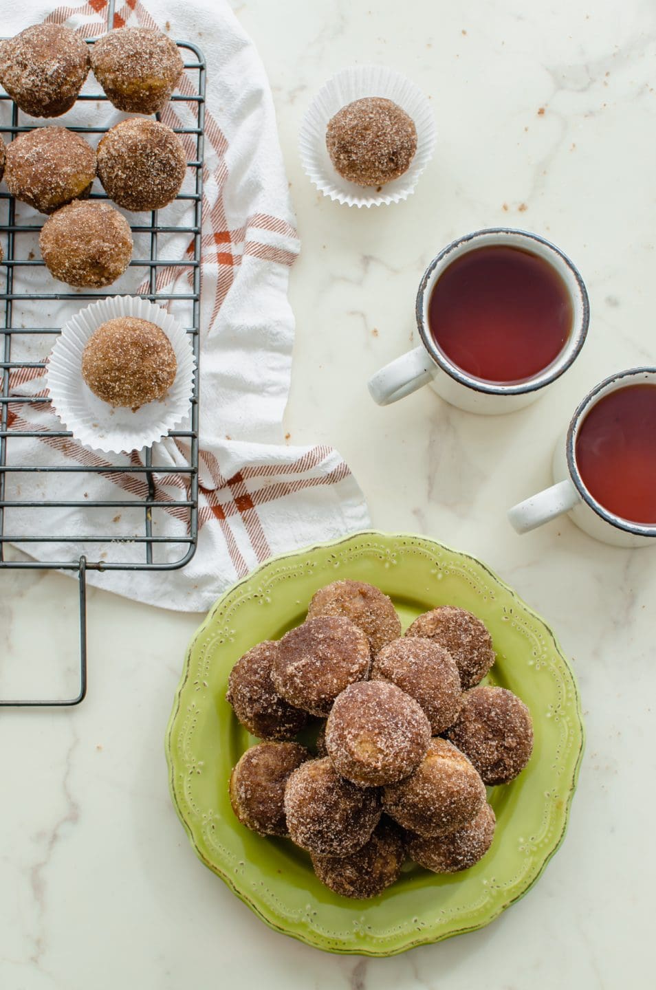 An overhead shot of apple cider donut holes on a green plate with cups of tea on the side.