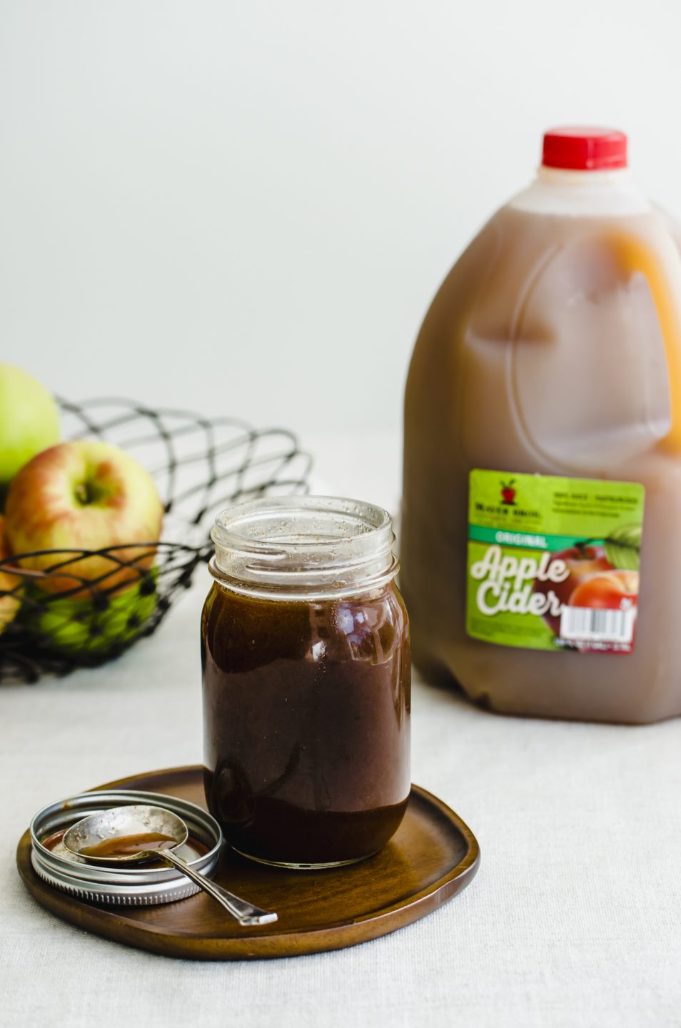 A jar of boiled apple cider syrup on a wooden plate with a jug of apple cider and a basket of fresh apples on the side. 