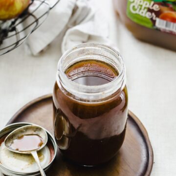 A jar of boiled apple cider syrup sitting on a wooden plate with a small spoon on the side.