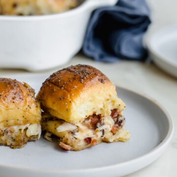 Two Kentucky Hot Brown Sliders on a blue ceramic plate with a baking dish of sliders and blue napkin in the background.