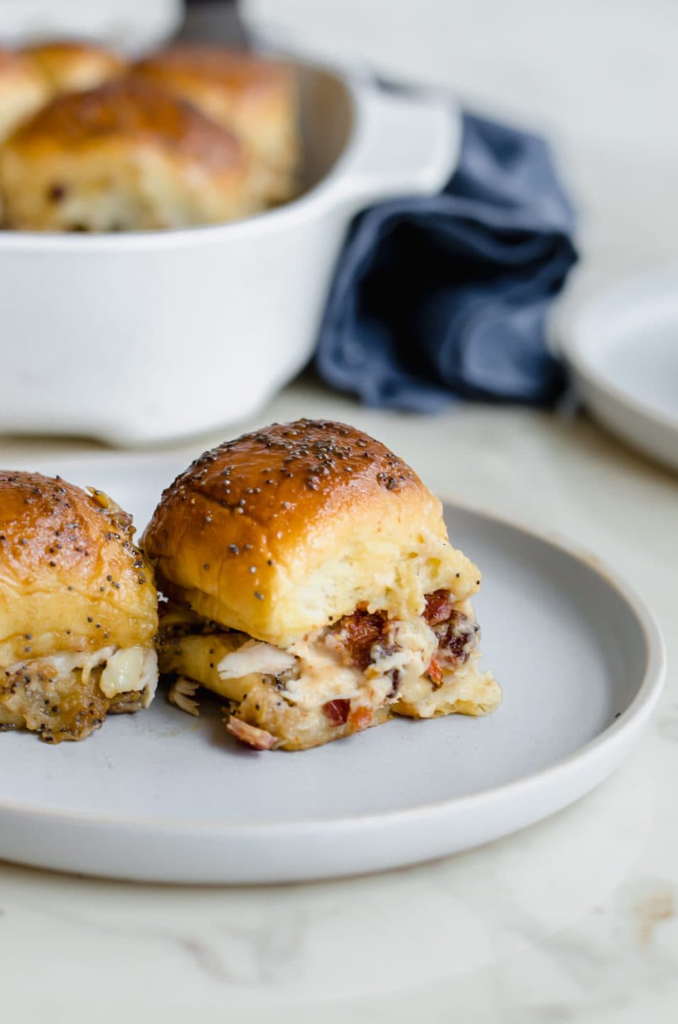 Two Kentucky Hot Brown Sliders on a blue ceramic plate with a baking dish of sliders and blue napkin in the background. 