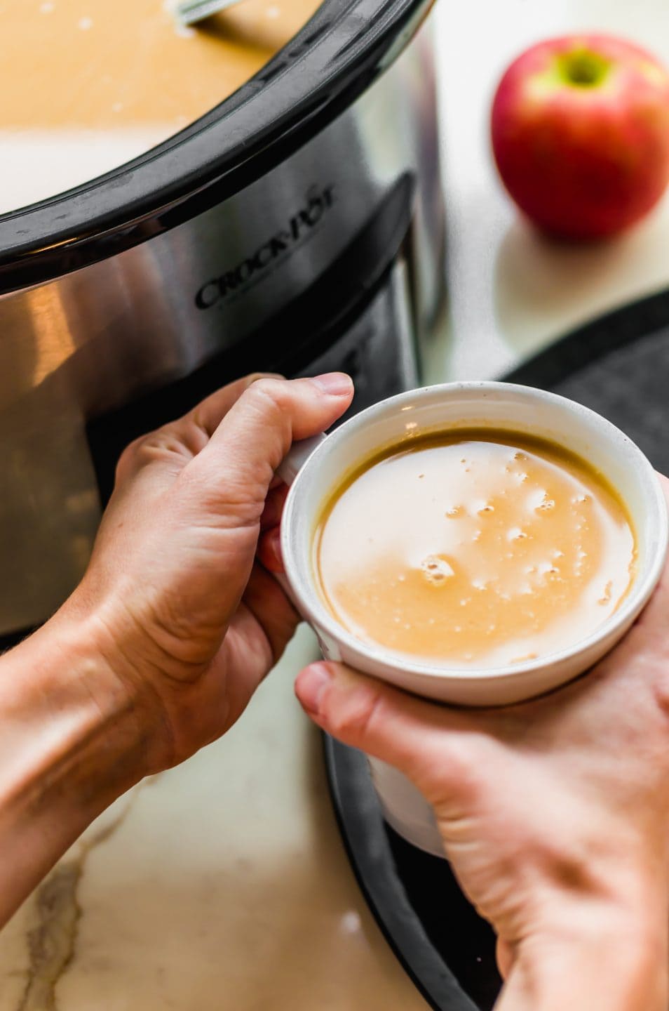 Two hands holding a white mug with salted caramel apple cider next to a Crock Pot.