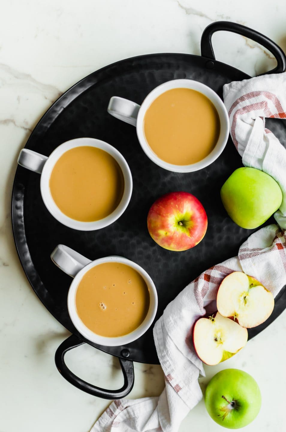 A black iron tray with three white mugs filled with salted caramel apple cider and apples on the side.