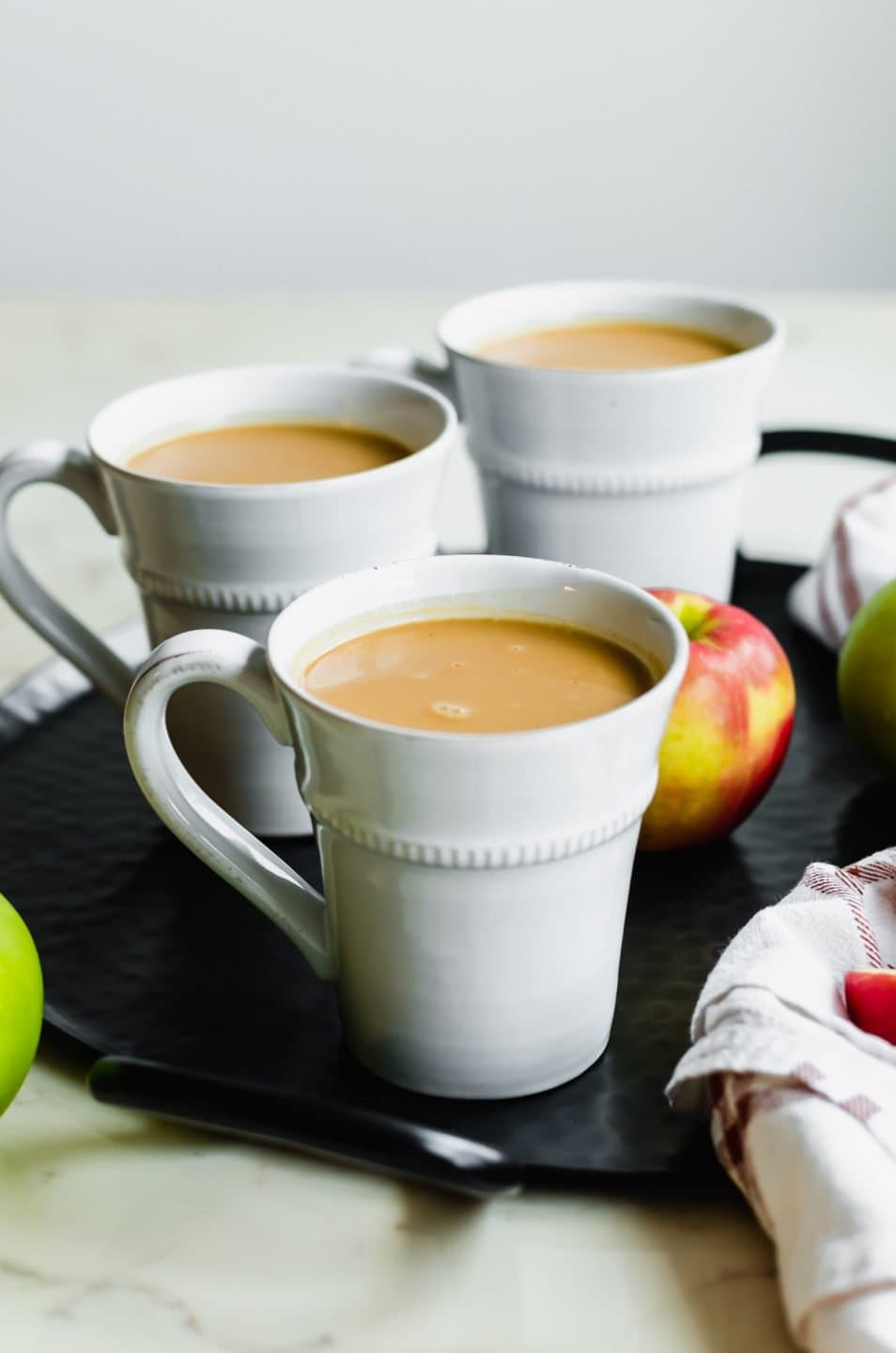 A black iron tray with three white mugs filled with salted caramel apple cider and apples on the side.