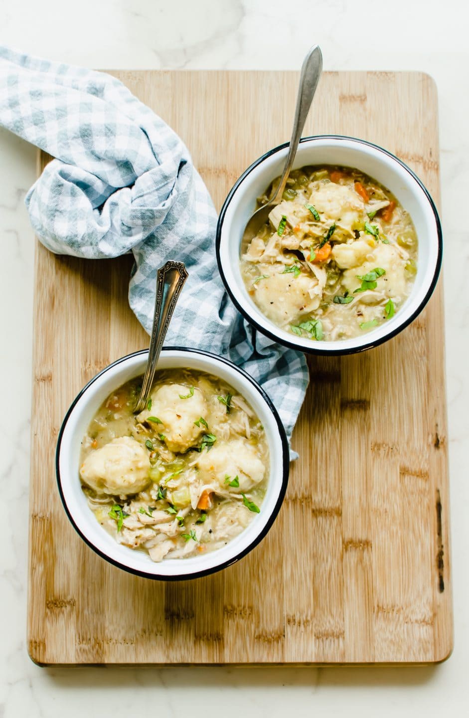 Two white enamel bowls of turkey and dumplings on a cutting board with a gingham dish cloth on the side.