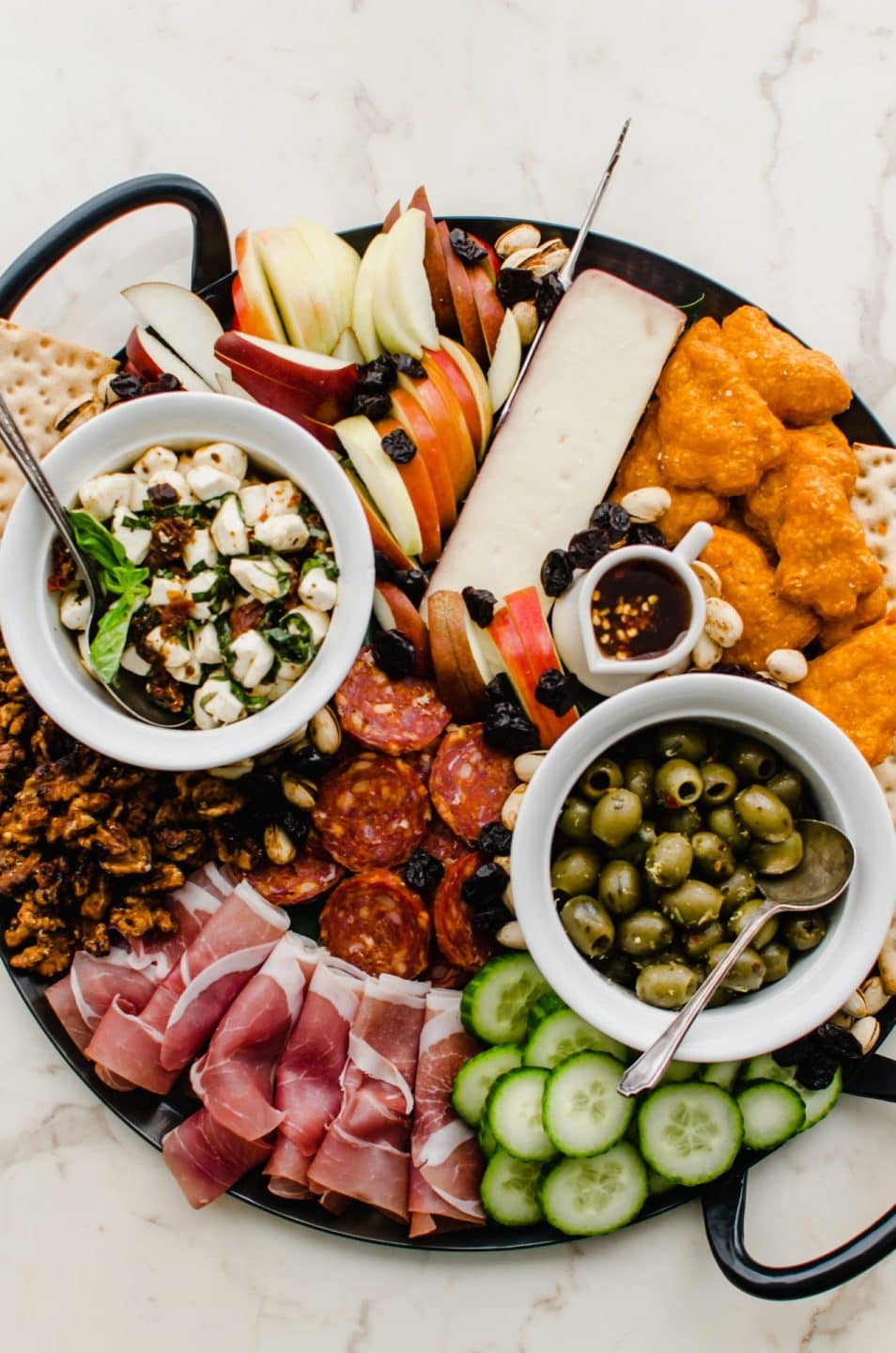 An overhead shot of a black iron platter filled with cheeses, olives, meats, spreads, fruit, and crackers. 