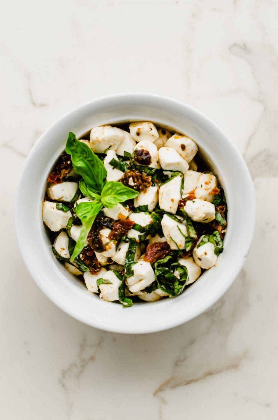 A white bowl filled with Italian marinated mozzarella pearls with a sprig of fresh basil on top.