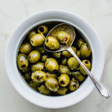 A white bowl filled with warm olives with lemon and rosemary with an antique spoon on the side.