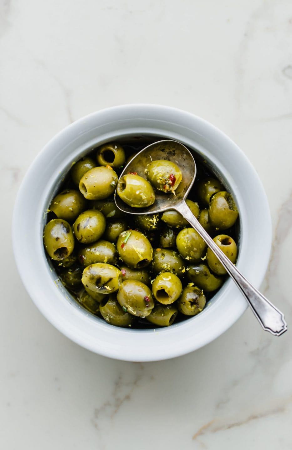 A white bowl filled with warm olives with lemon and rosemary with an antique spoon on the side. 