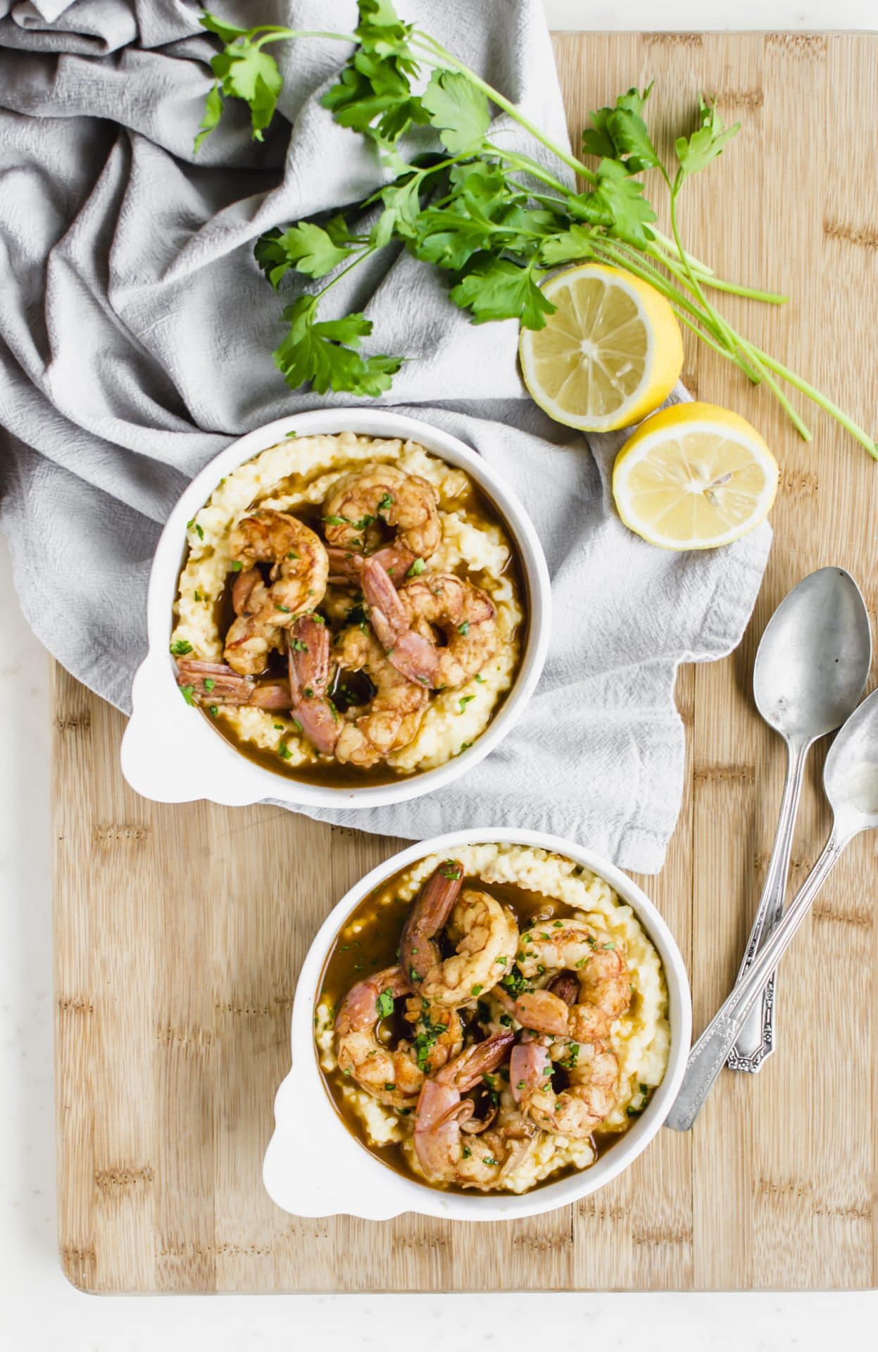 An overhead shot of two bowls of barbecue shrimp and grits on a cutting board with a lemon on the side. 