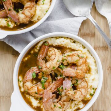 A close up shot of a white bowl filled with barbecue shrimp and grits with an antique spoon on the side.