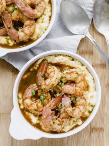 A close up shot of a white bowl filled with barbecue shrimp and grits with an antique spoon on the side.