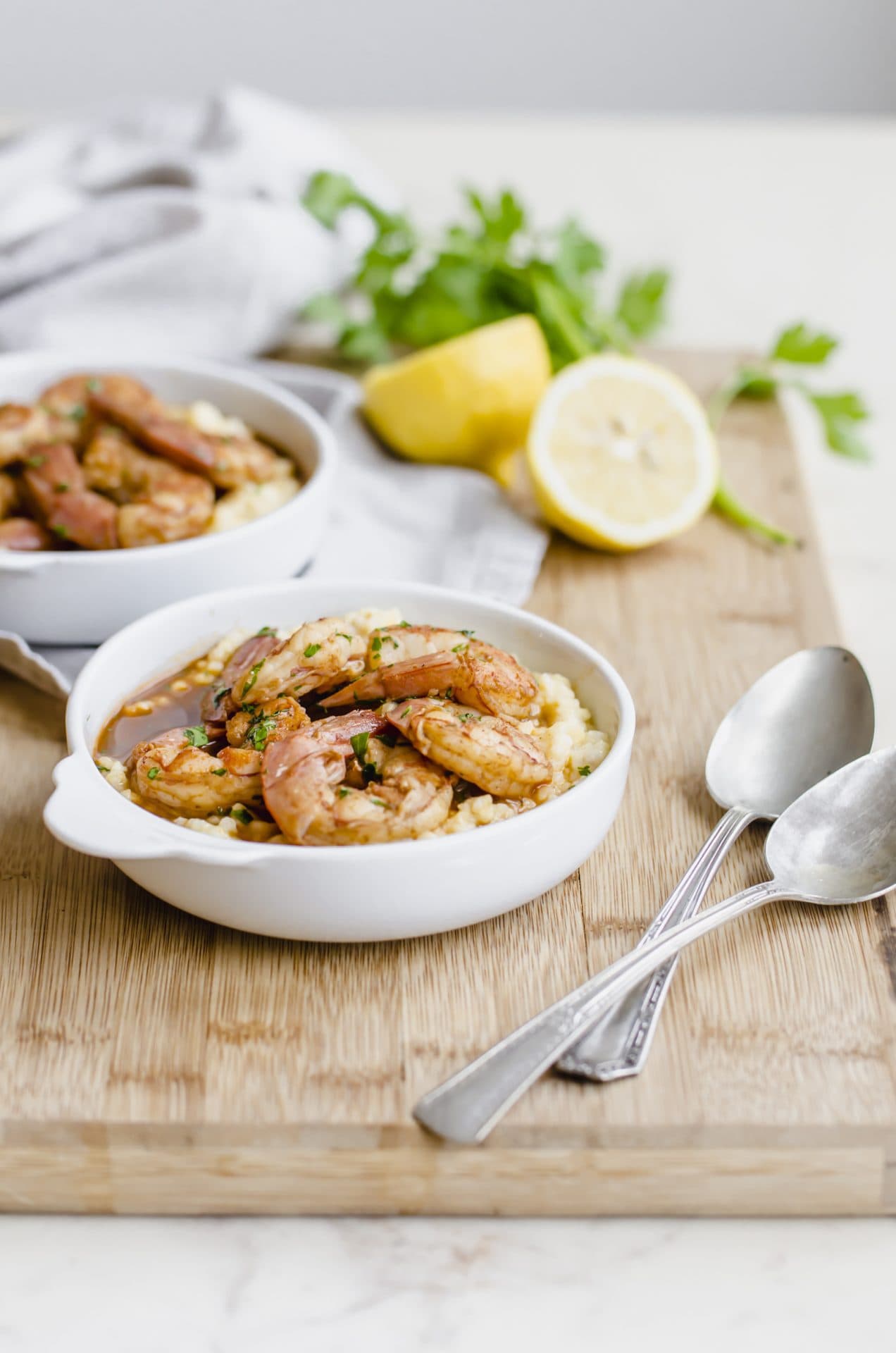 An overhead shot of two bowls of barbecue shrimp and grits on a cutting board with a lemon on the side.