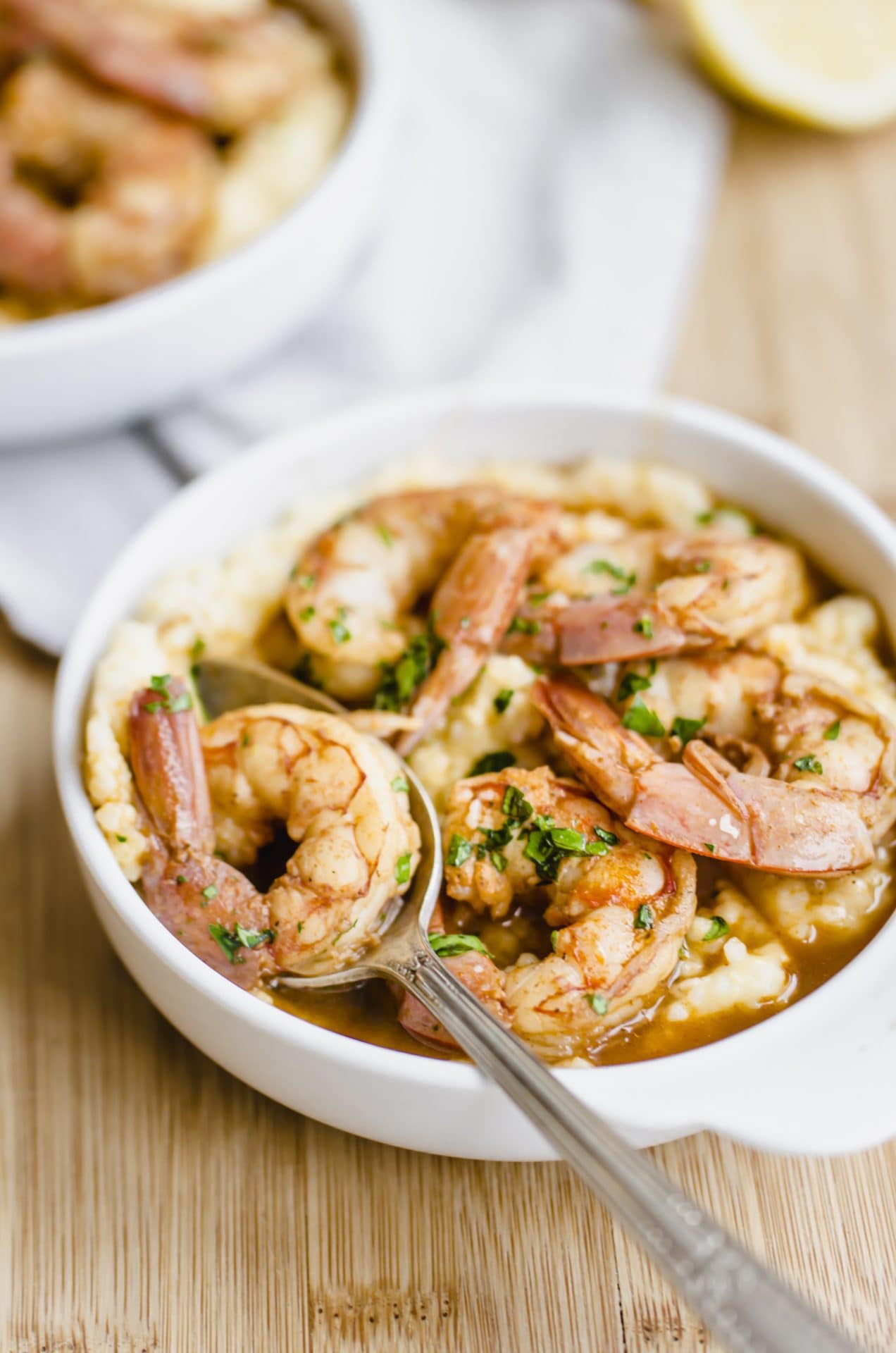 A close up shot of a white bowl filled with barbecue shrimp and grits with an antique spoon on the side.