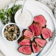 A white round platter with sliced beef tenderloin ad dishes of horseradish cream and marsala mushrooms.