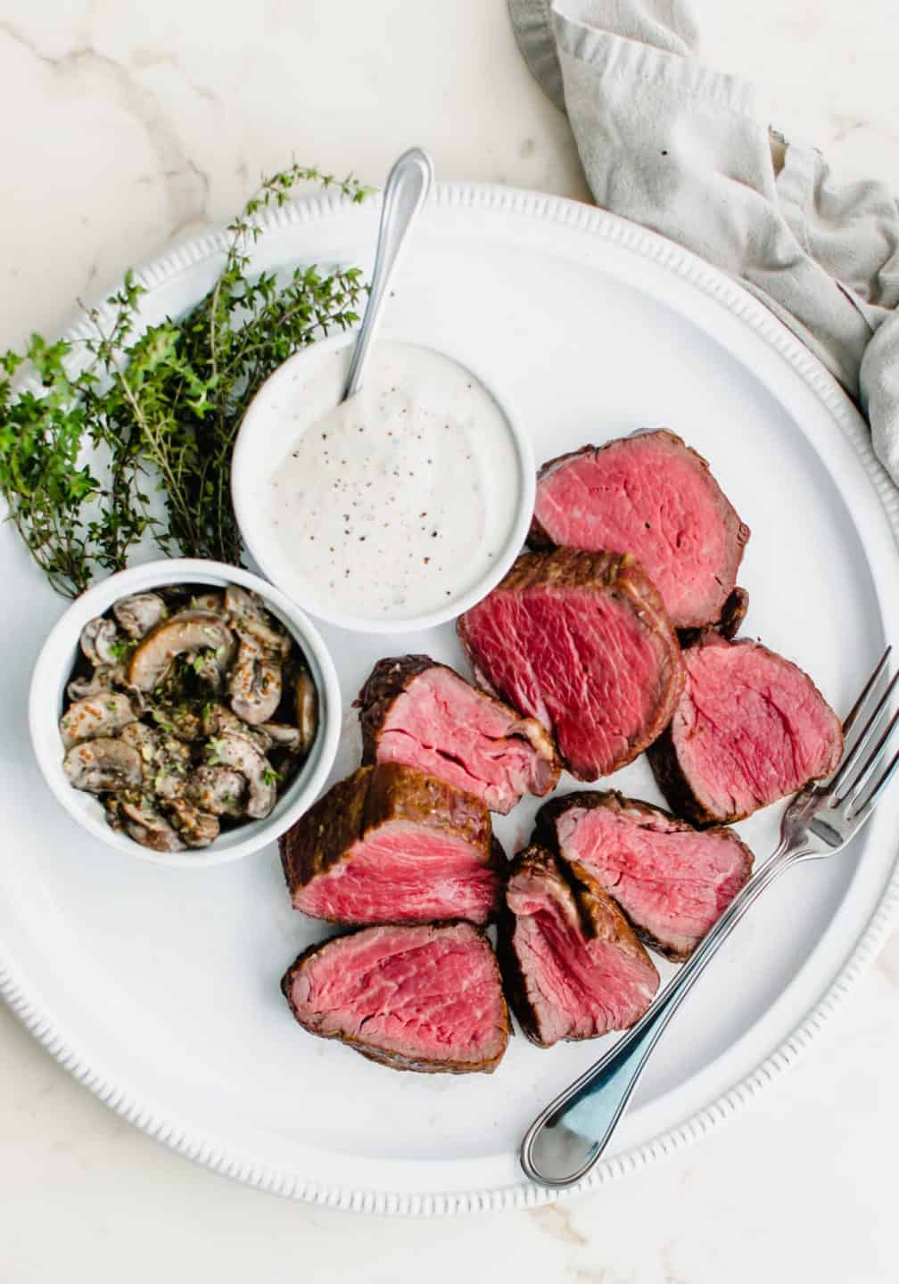 A white round platter with sliced beef tenderloin ad dishes of horseradish cream and marsala mushrooms.