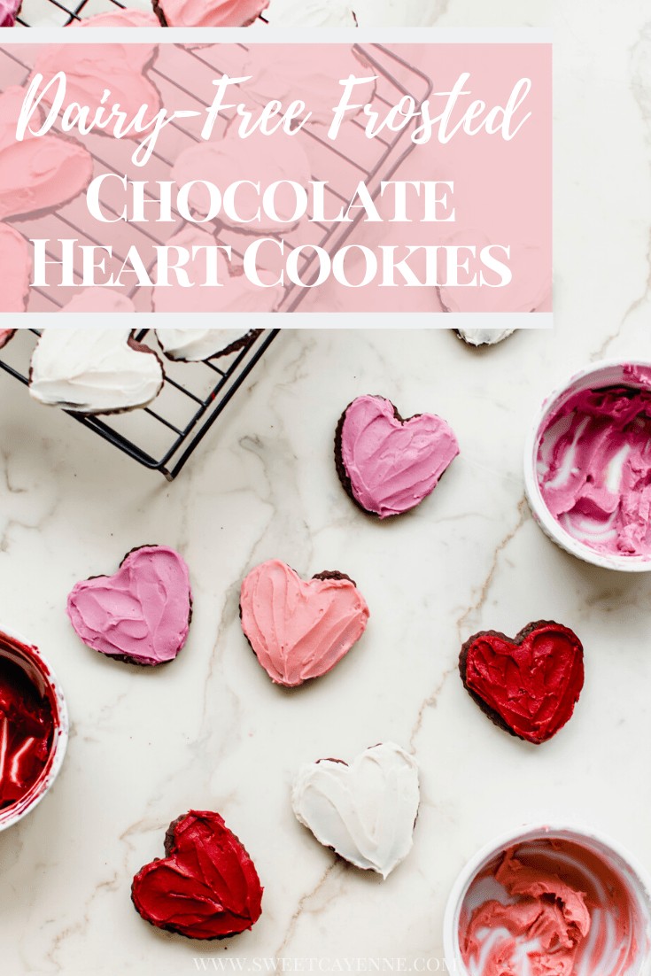 Frosted heart chocolate cutout cookies on a white countertop with bowls of frosting on the side.