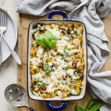 A blue casserole dish with Chicken Rotini Bake on a cutting board with a dish towel and serving spoon on the side.