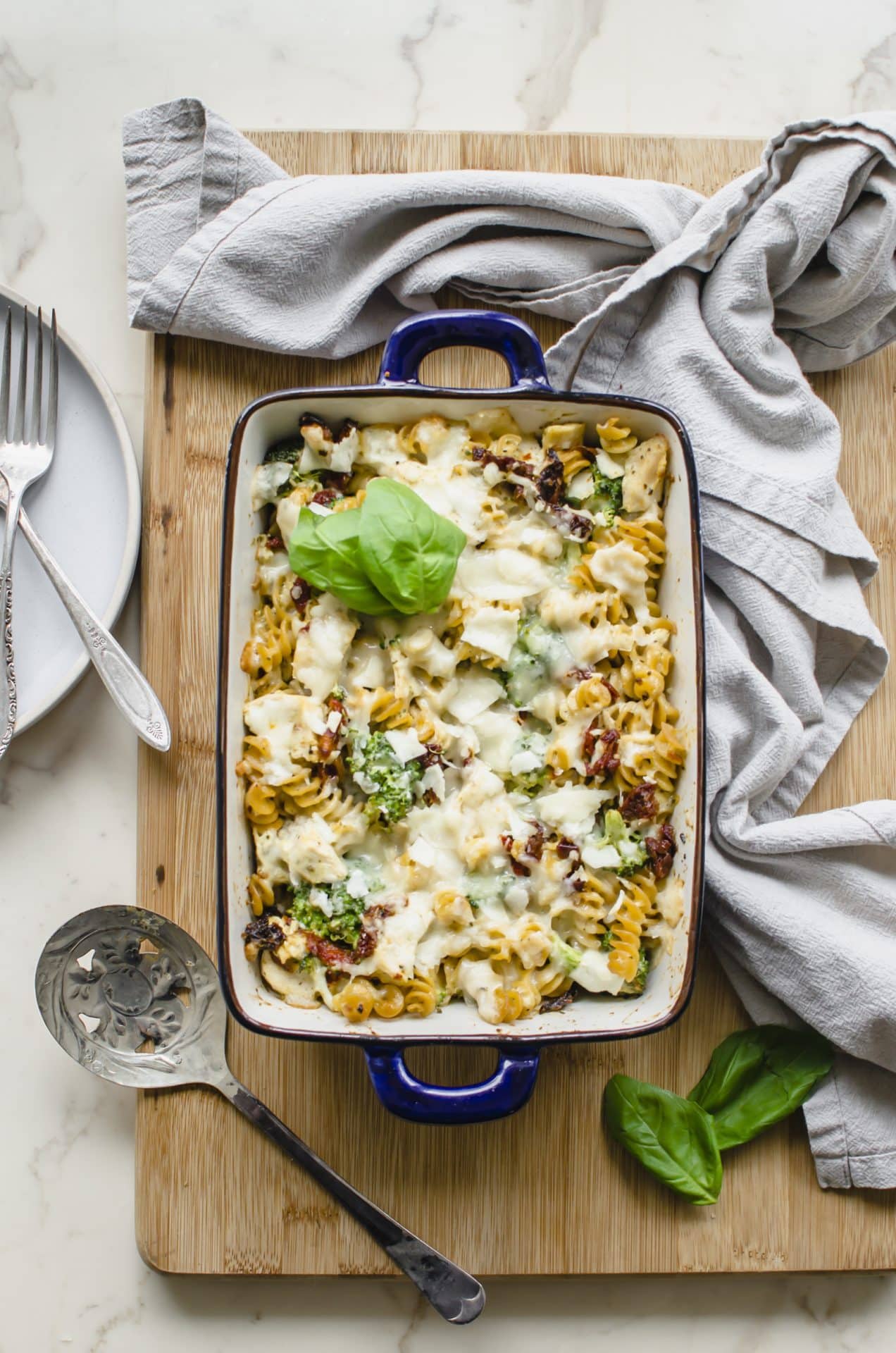 A blue casserole dish with Chicken Rotini Bake on a cutting board with a dish towel and serving spoon on the side. 