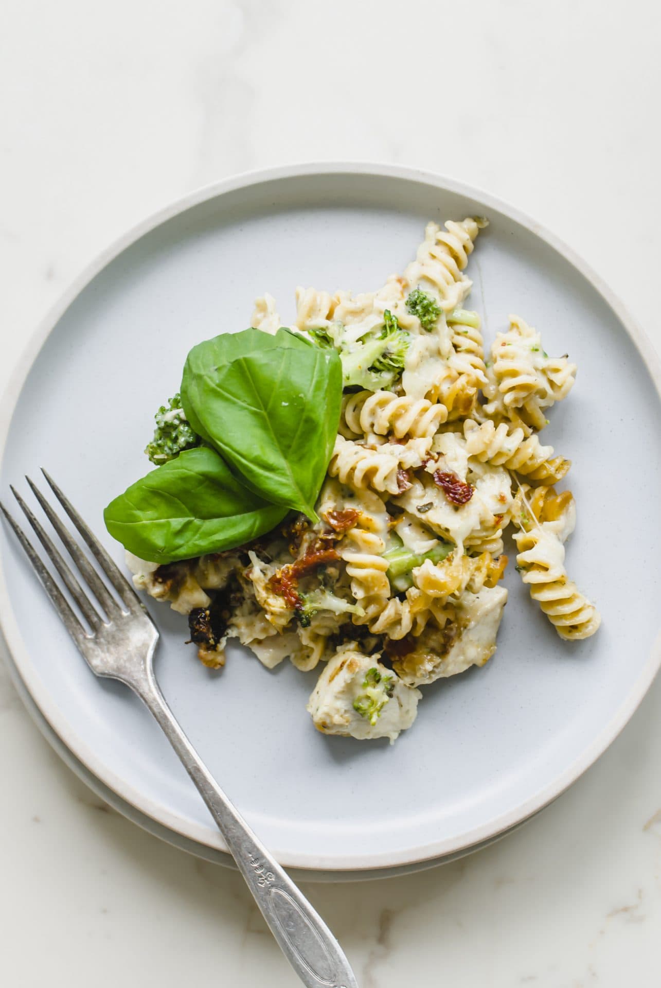 A blue plate with a serving of chicken rotini bake with a sprig of basil on top and a fork on the side.