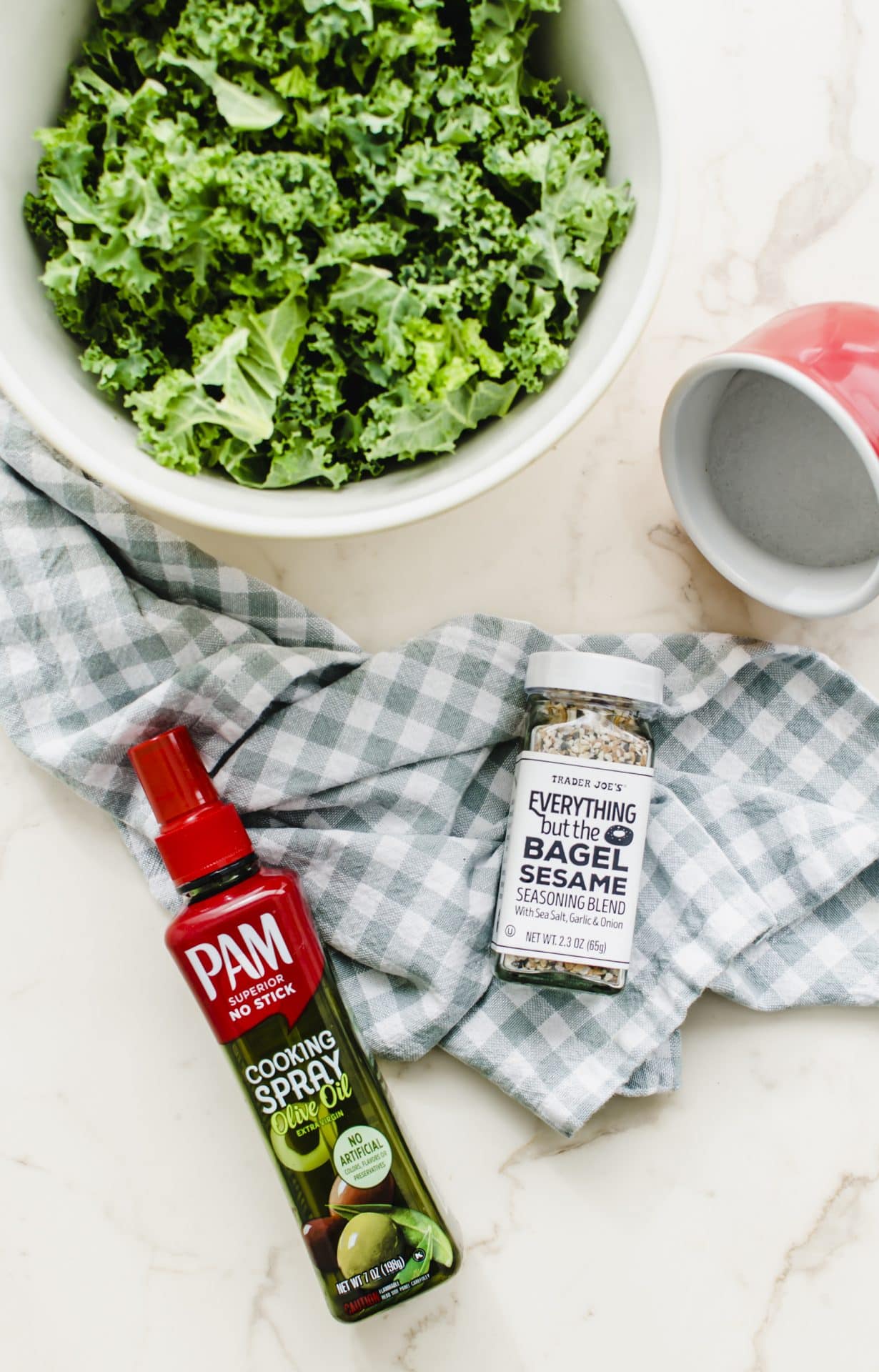 A bottle of olive oil spray, a salt holder, a bowl of kale leaves and a bottle of Everything bagel seasoning on a white counter.