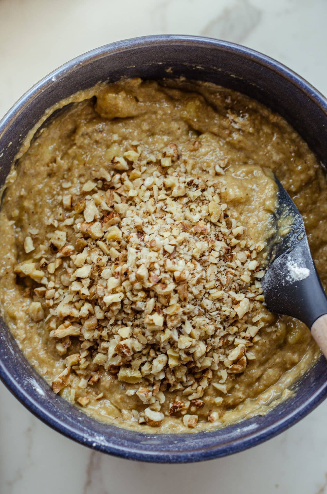 A gray rubber spatula folding walnuts into a banana bread batter in a blue bowl. 