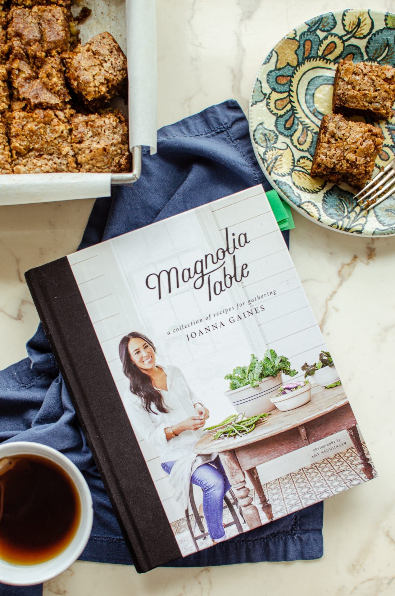 The Magnolia Table cookbook on a blue napkin with a plate of banana bread and a white cup of tea on the side. 