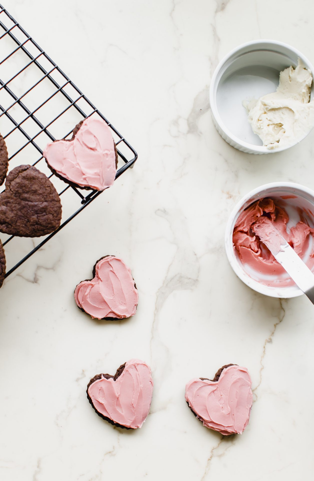Frosted heart chocolate cutout cookies on a white countertop with bowls of frosting on the side.